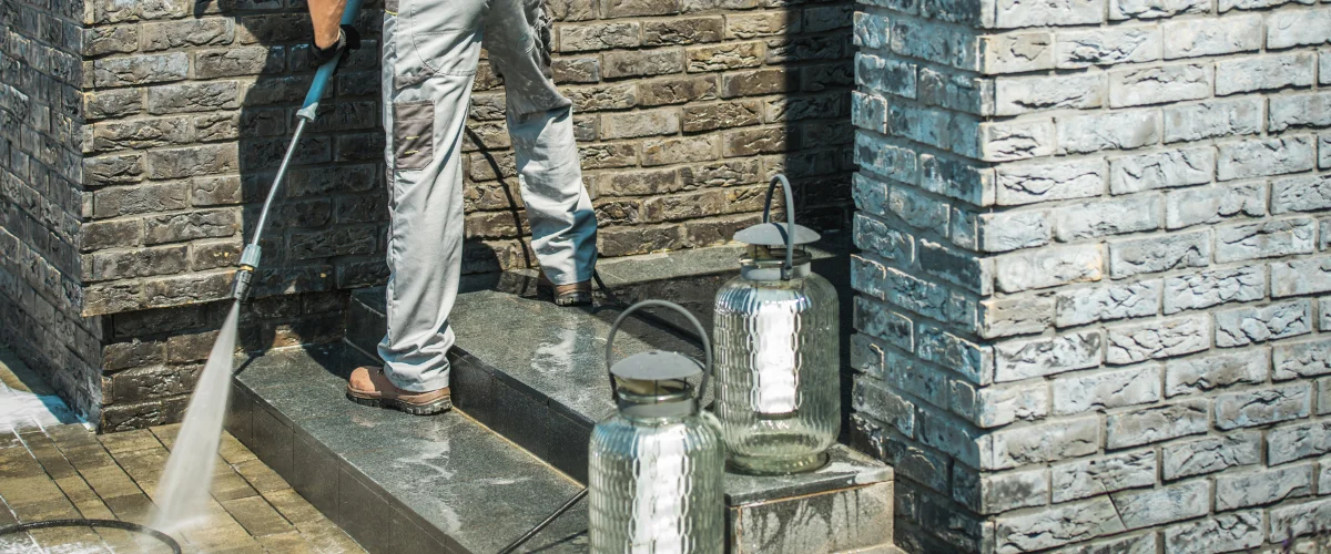 A person in light pants power washing a dark tiled patio next to a stone wall, with two glass lanterns nearby.