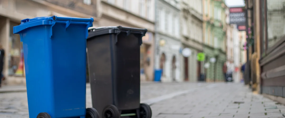 A tall, weathered blue plastic trash can with wheels stands on a paved surface next to green grass and foliage. A tall, weathered blue plastic trash can with wheels stands on a paved surface next to green grass and foliage.