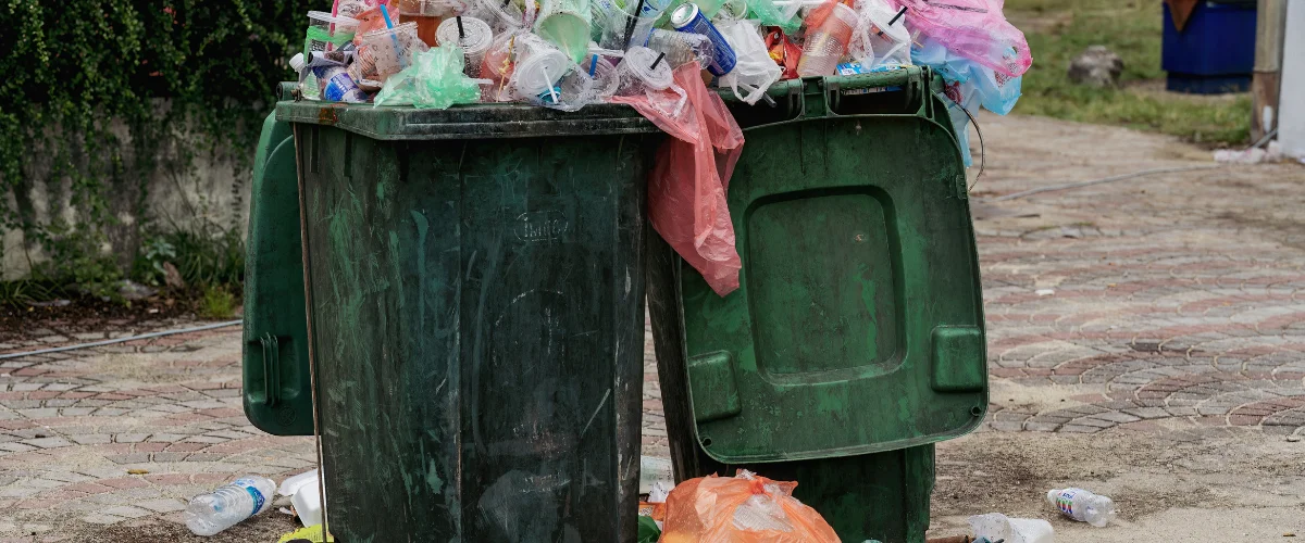 A dark green dumpster filled to overflowing with plastic waste, bags, and cups sits on a brick path. A dark green dumpster filled to overflowing with plastic waste, bags, and cups sits on a brick path.