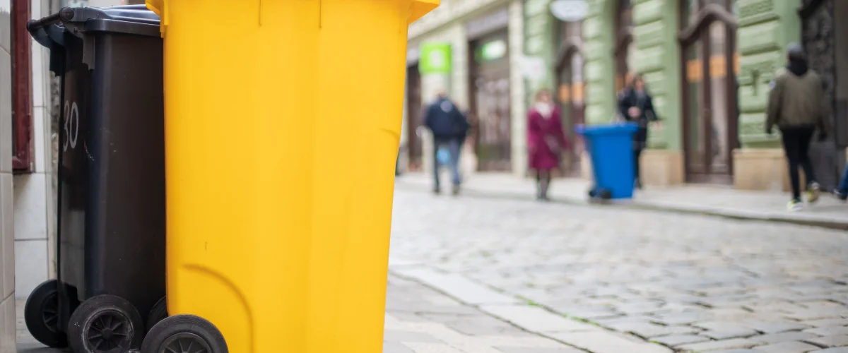 A black and a bright yellow trash bin with wheels on a cobblestone street with blurred people walking in the background. A black and a bright yellow trash bin with wheels on a cobblestone street with blurred people walking in the background.