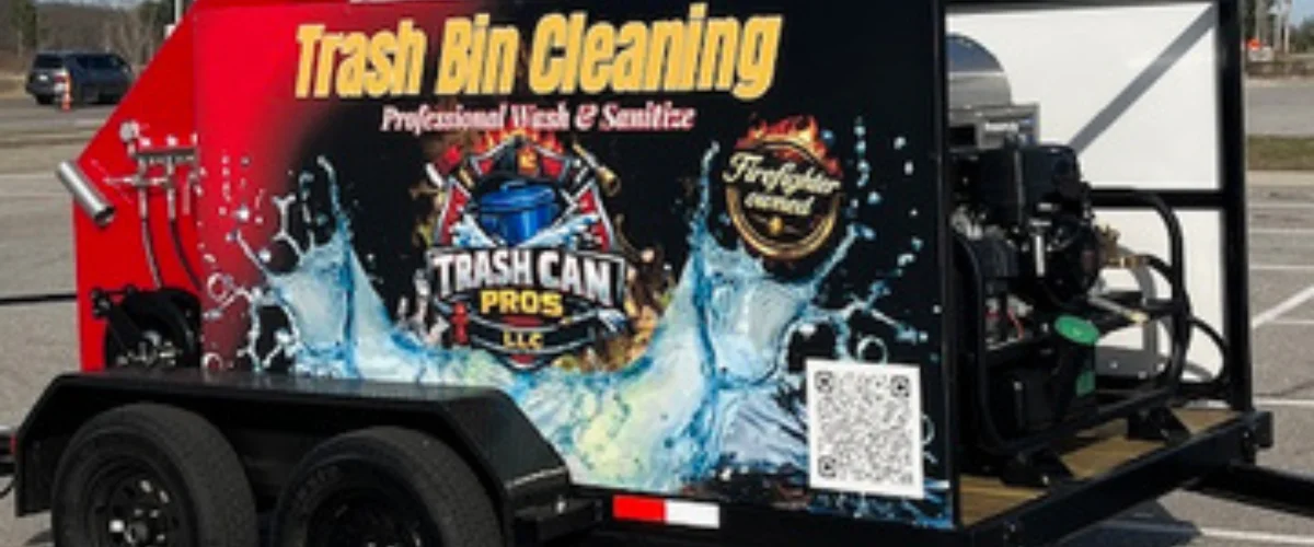 A bright red trailer with "Trash Bin Cleaning" branding and a green WM trash bin tilted inside its cleaning mechanism.