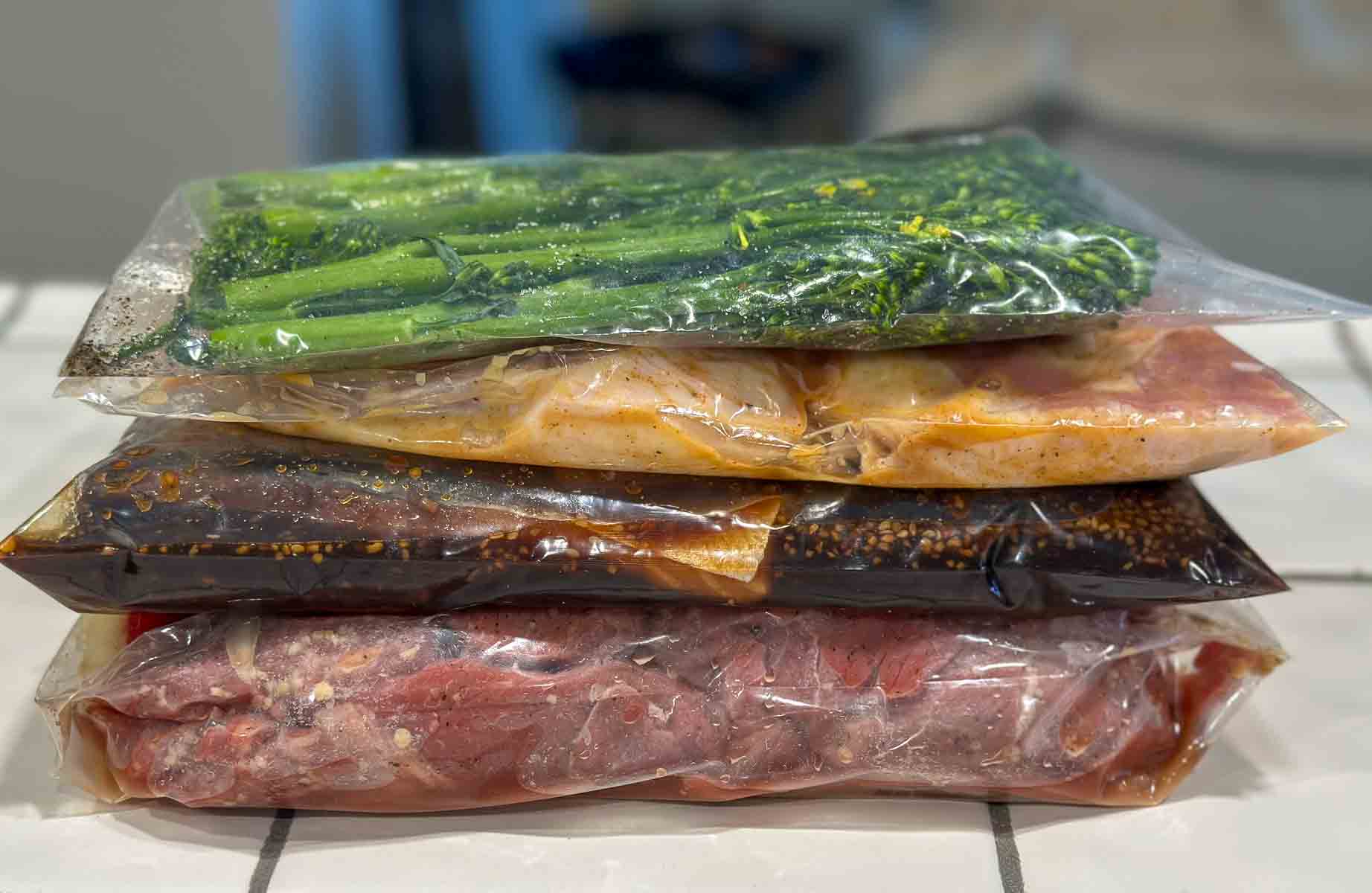Stack of sealed freezer bags filled with prepped dinner ingredients like broccoli, chicken, sauces, and beef. Informative, product-related.