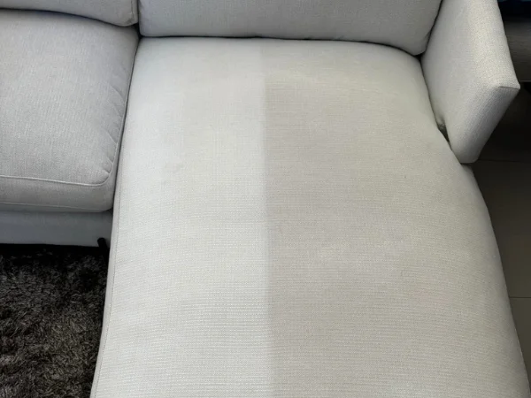 A professional cleaner using a heavy-duty industrial steam cleaning machine to deep-clean a tan fabric sofa in a well-lit living room.