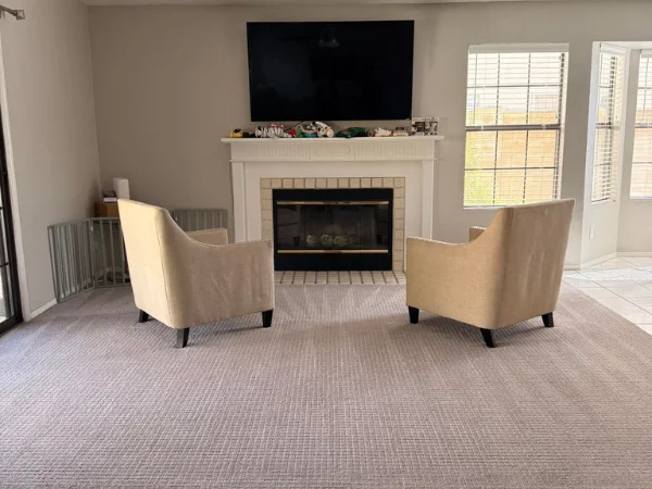 A living room featuring two beige armchairs facing a white fireplace with a TV mounted above, and windows with blinds.