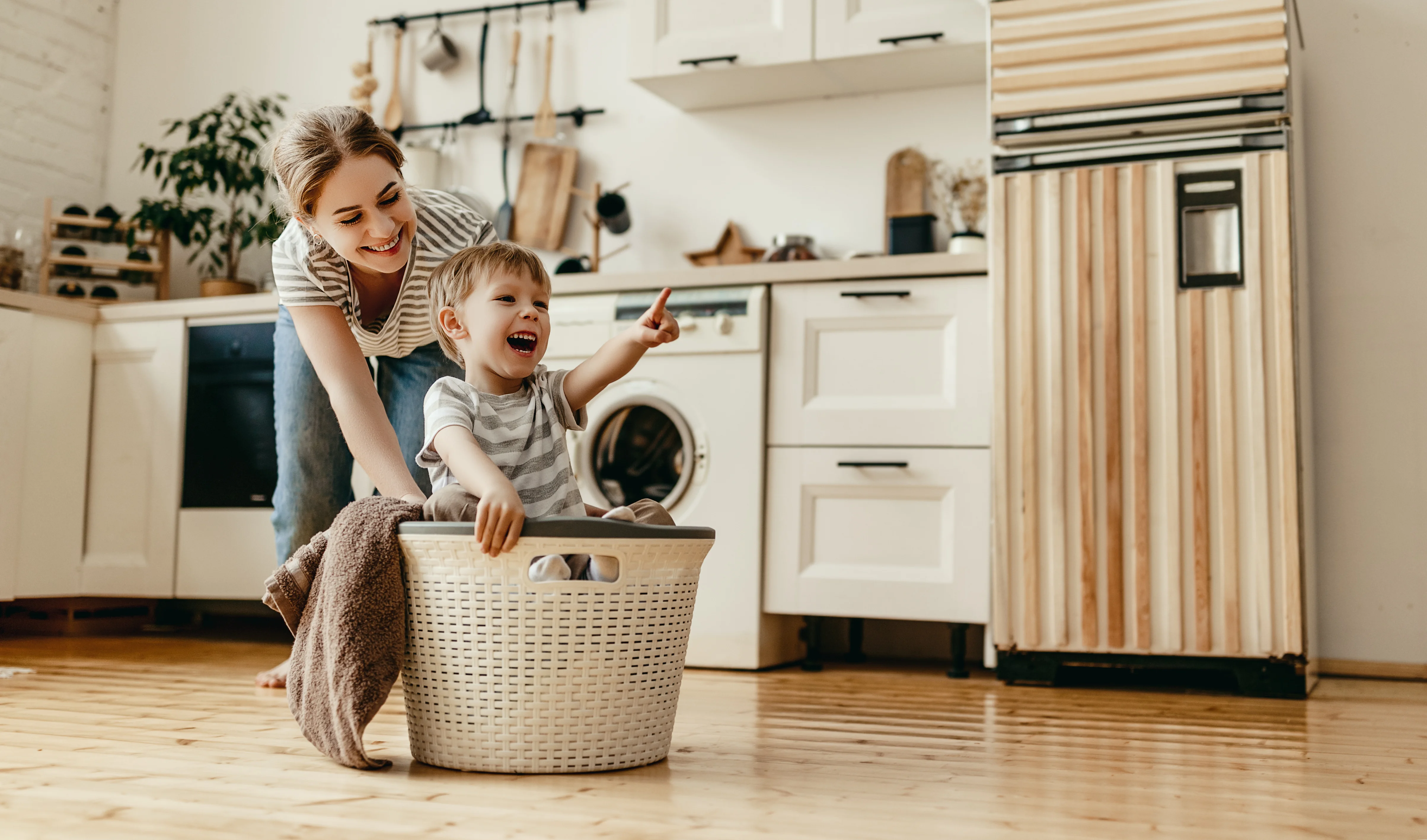 A happy mother smiles at her young son playing in a laundry basket on the kitchen floor.