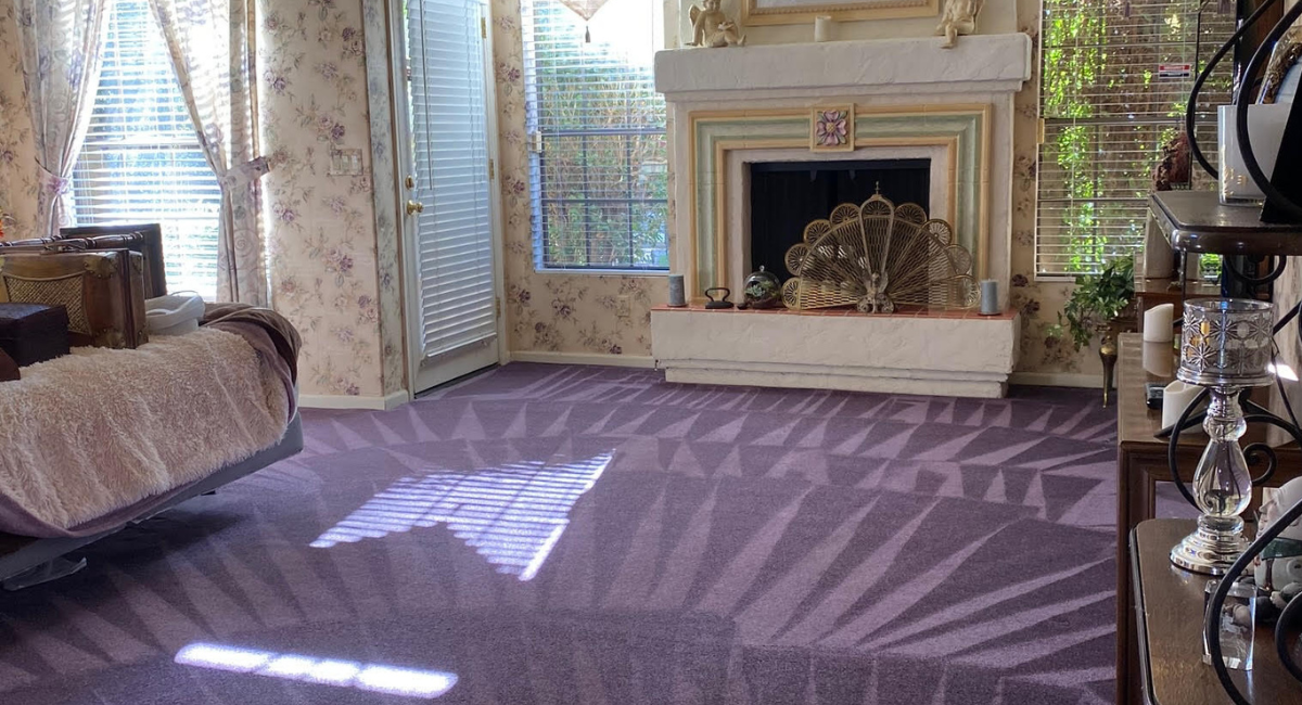 A living room with light purple floral wallpaper, purple patterned carpet, and a white fireplace with a peacock screen. A living room with light purple floral wallpaper, purple patterned carpet, and a white fireplace with a peacock screen.