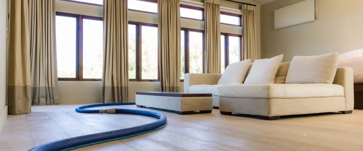 A blue hose lies across a light wood floor in a living room with large windows and an off-white sofa.