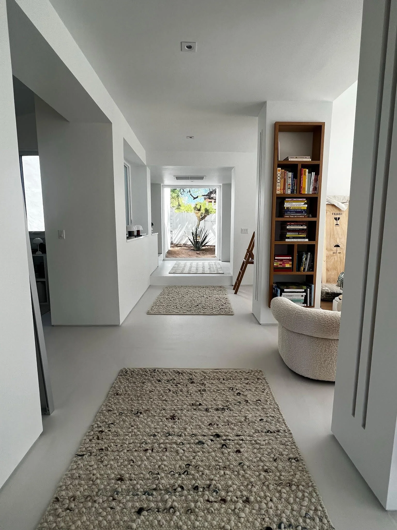 A minimalist white hallway with two textured rugs leading to an outdoor desert garden, with a tall bookshelf and armchair.