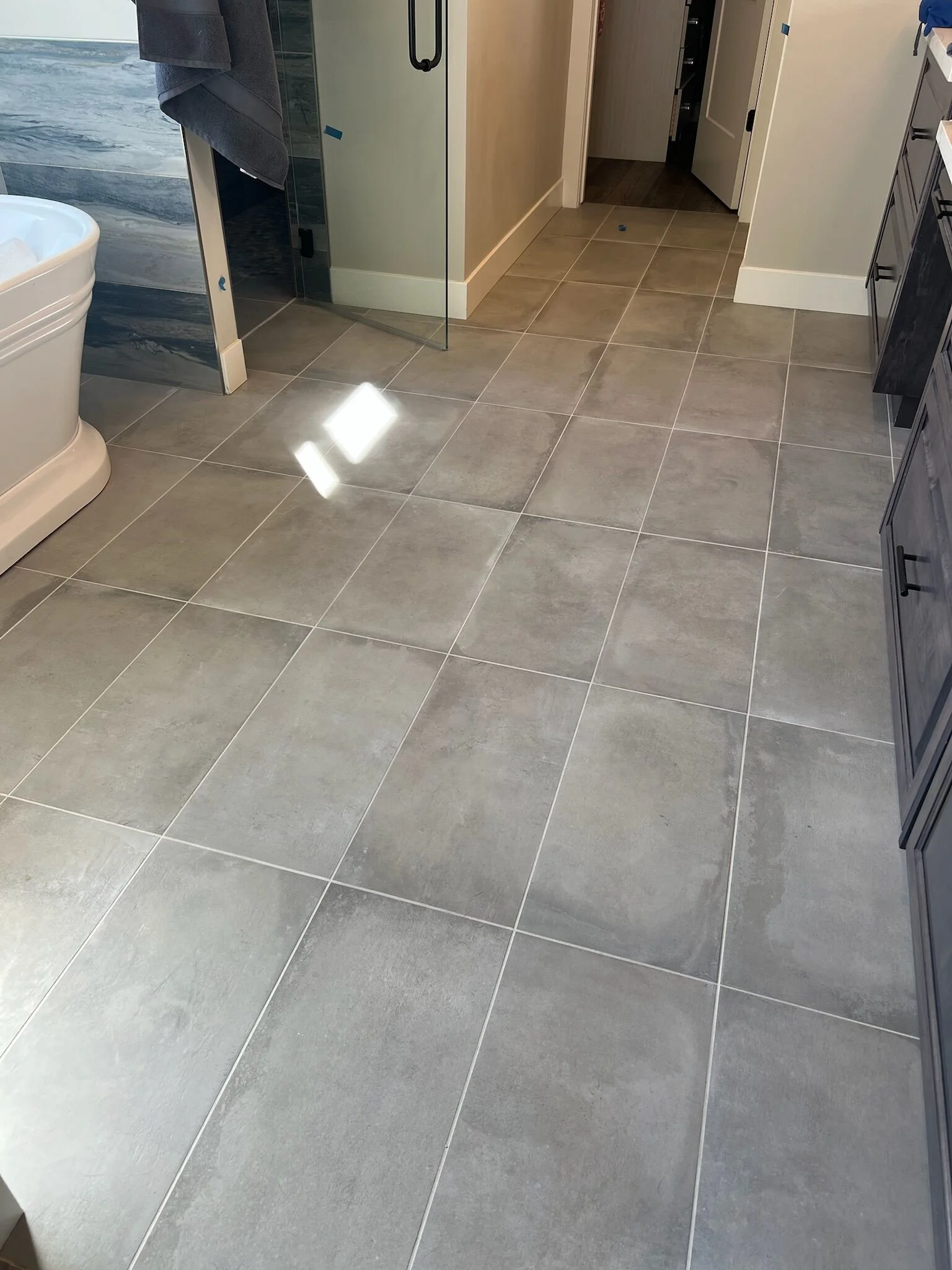 Wide view of a modern bathroom with light gray tiled flooring, a white freestanding tub, and a dark wood vanity.