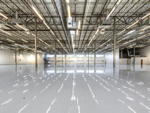 A wide, empty industrial hall with a reflective light grey floor and exposed ceiling trusses and lights.
