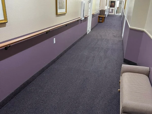 A long, carpeted hallway with a light purple lower wall, light beige upper wall, and a wooden handrail.