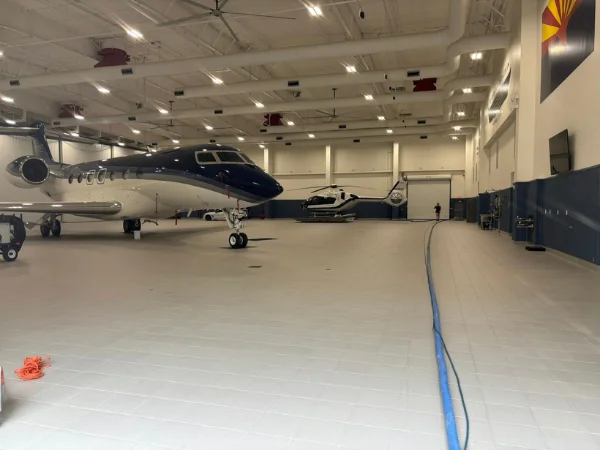 A luxurious private jet and a helicopter parked inside a brightly lit aerospace hangar with a blue hose on the floor.