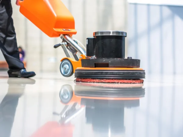 A close-up of a person operating an orange industrial floor buffer over a shiny, reflective floor, with the machine's reflection clearly visible.