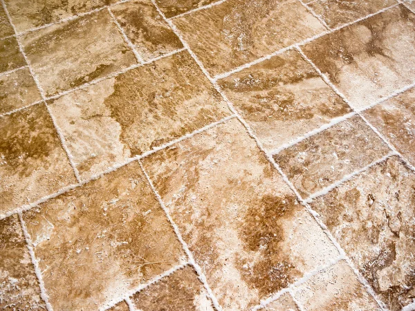 A close-up, high-angle view of a tiled floor made of varied tan and brown stone with rough white grout.