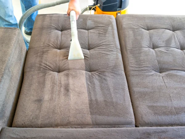 A person uses a cleaning tool to shampoo and clean a brown sofa, showing a visible clean and dirty split.