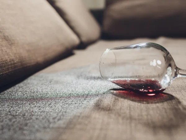 A wine glass lies spilled on a textured brown sofa, with red wine staining the fabric.