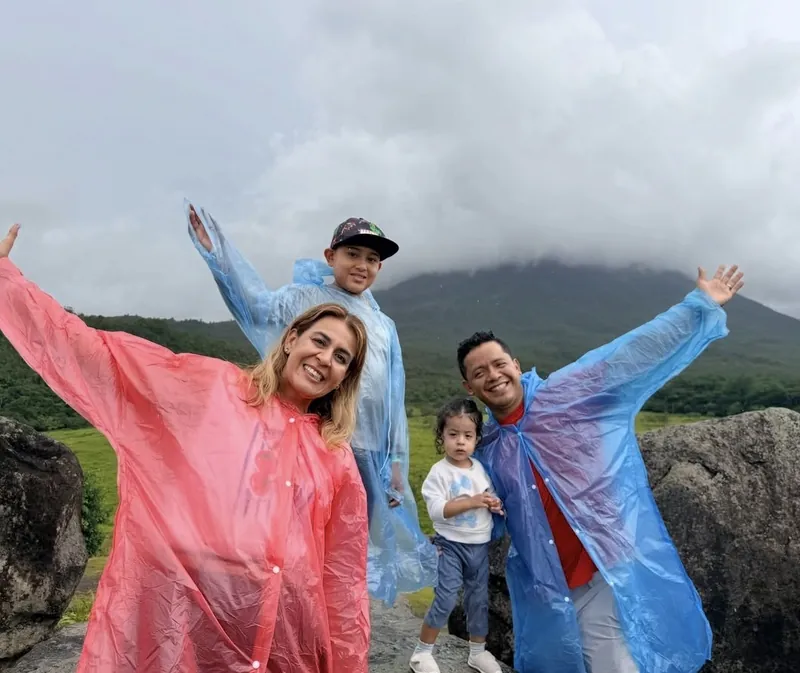 Alma Sández de viaje con su esposo y sus hijos, con impermeables y los brazos arriba frente a un volcán