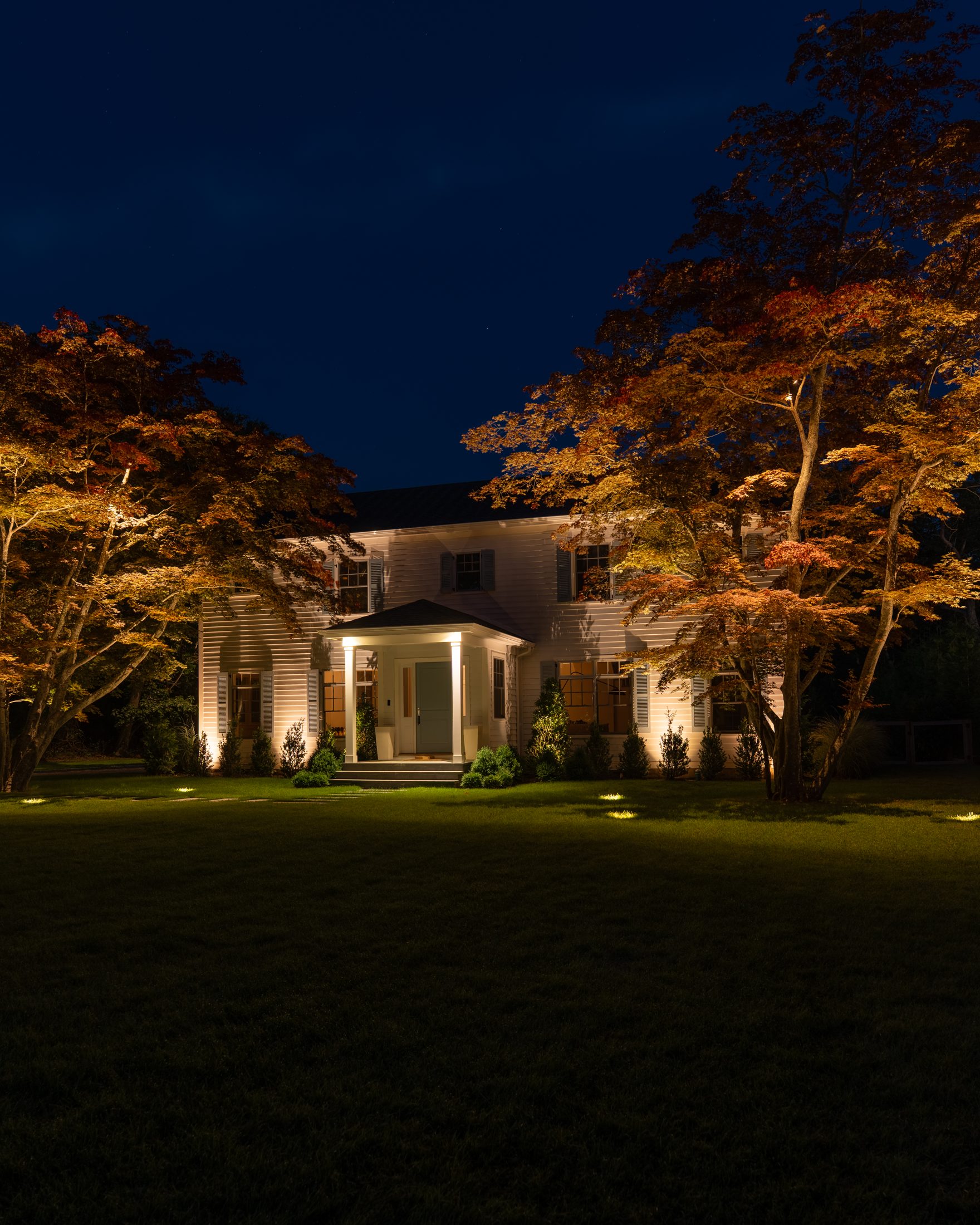 White clapboard house at night with Japanese maples uplit in warm light