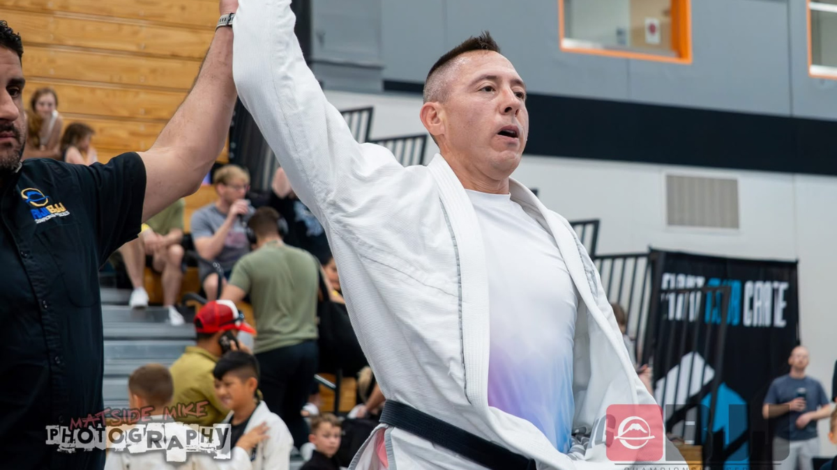 A jiu jitsu competitor in a white gi has his arm raised by the referee after a match.