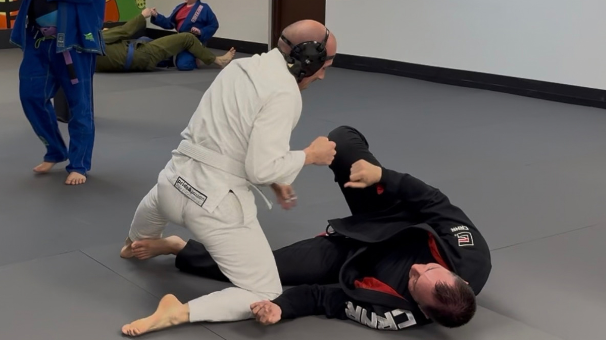 A training partner in a black gi makes a shaka gesture while lying beside a kneeling partner in a white gi on the mats.