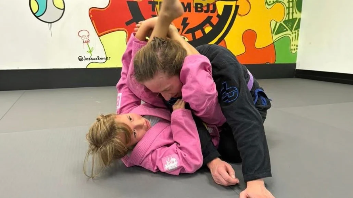 Two jiu jitsu practitioners in pink and black gis working through a guard position on the mats.