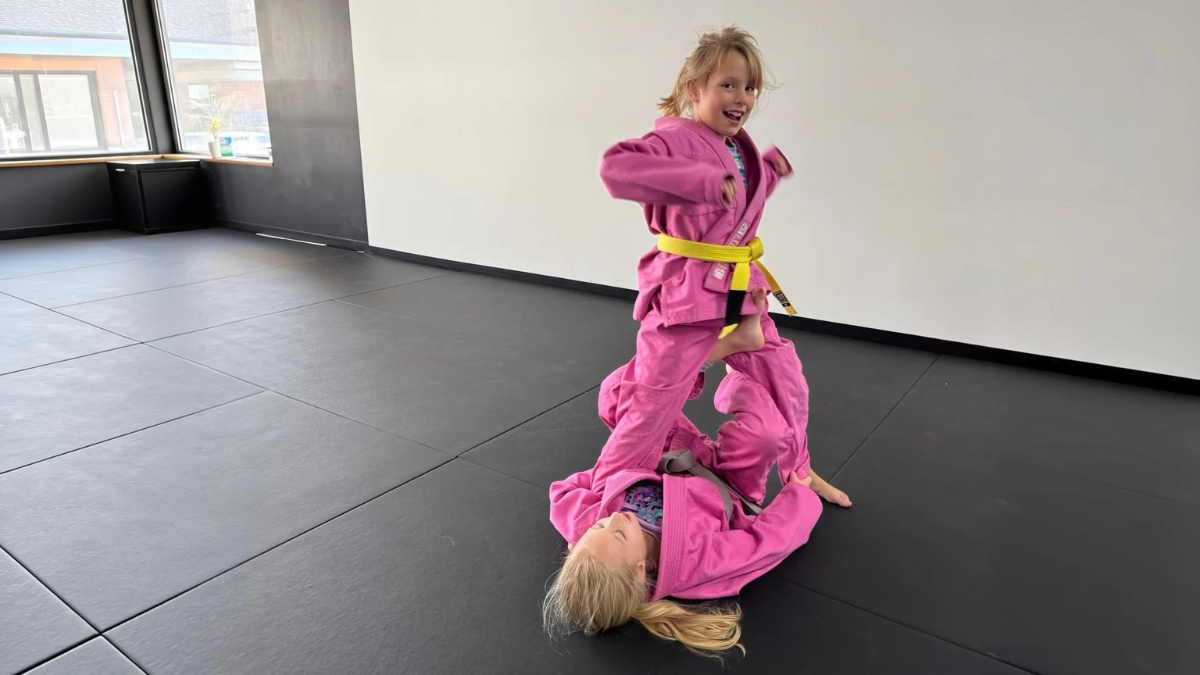 Two children in pink gis practicing jiu jitsu together on the mats, one standing and one on her back.