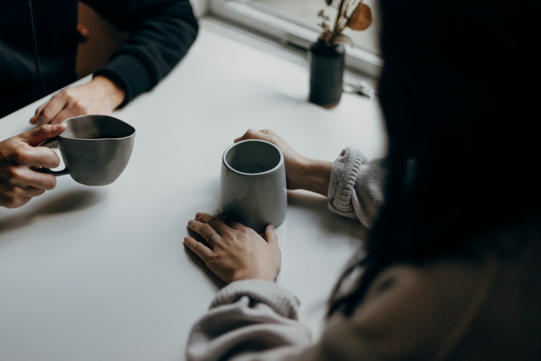 Two people seated at a pale table holding grey mugs, hands and sleeves visible, faces out of frame, soft window light.
