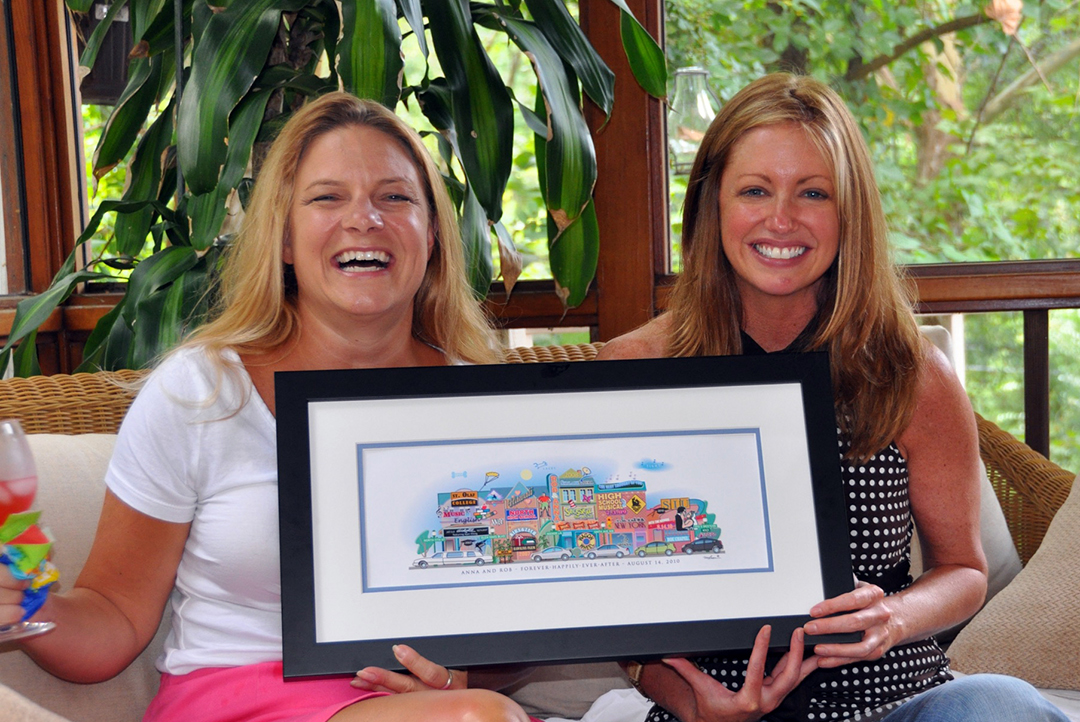 Two women holding a framed Your Memory Lane 5-building wedding street print.