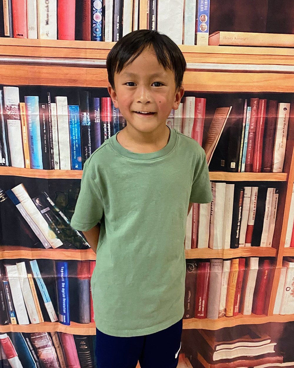 A boy in a green t-shirt smiling in front of a bookshelf backdrop on the first day of school.