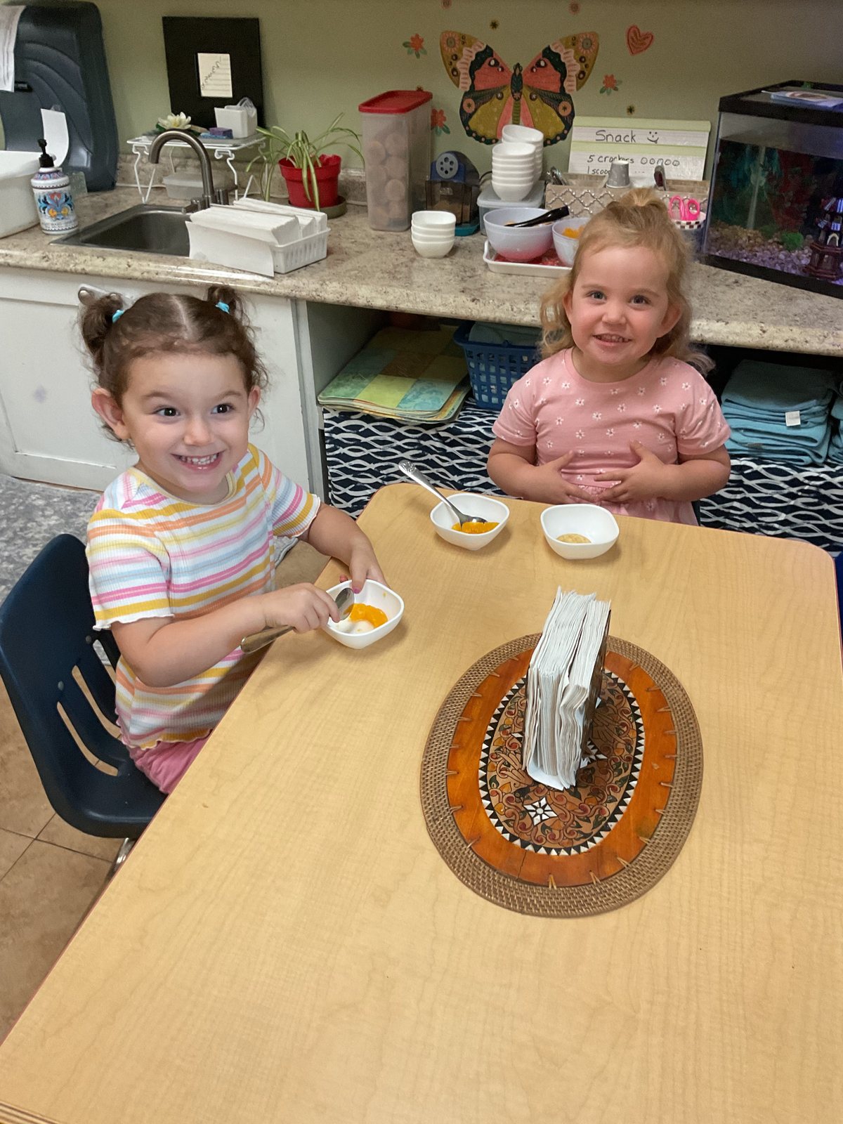 Two girls at a table doing a spooning exercise with small bowls, one smiling at the camera, a kitchen counter and butterfly wall art behind.