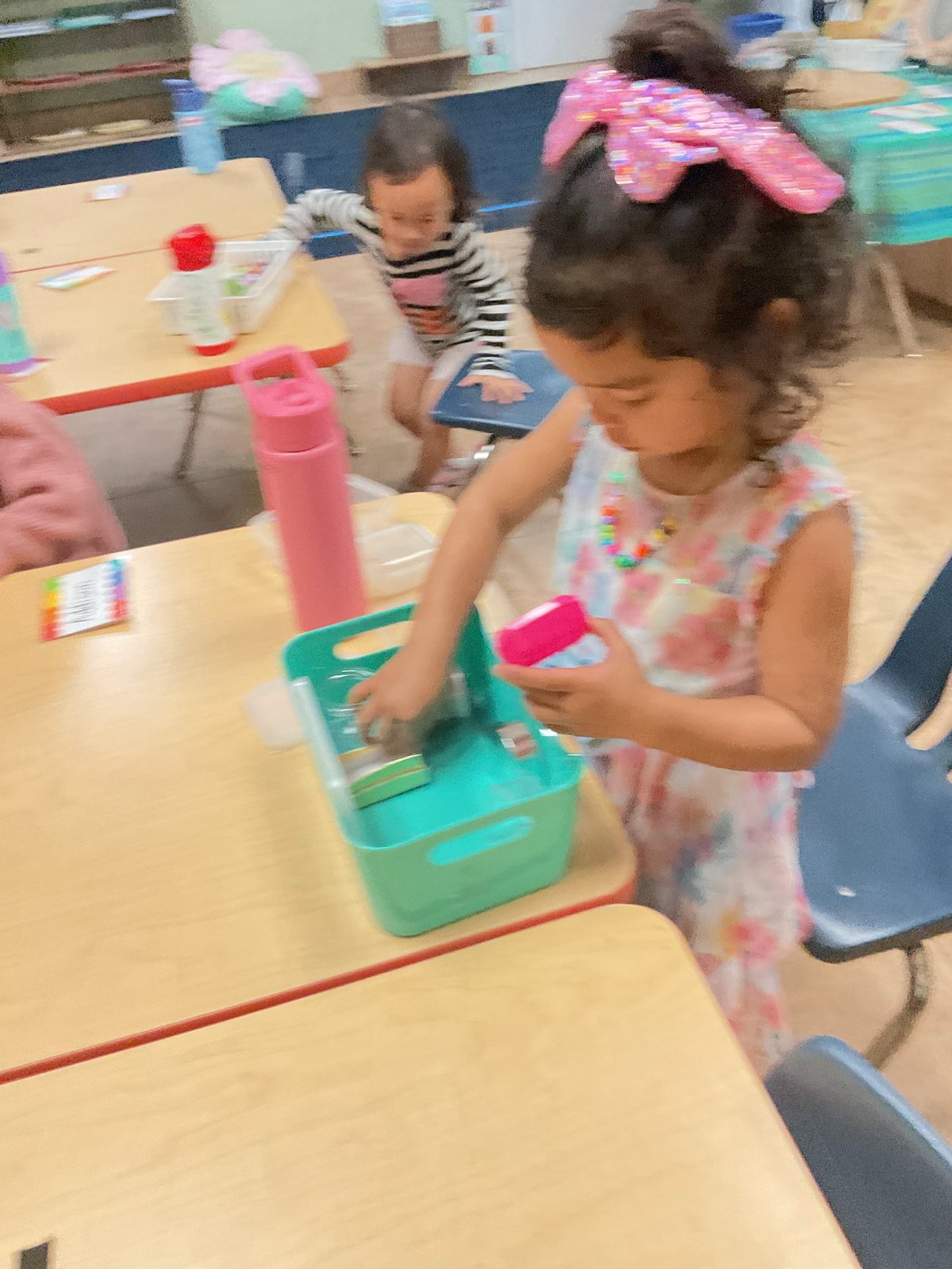 A girl in a floral dress with a pink hair bow carefully pouring at a table with a teal tray, another child working in the background.