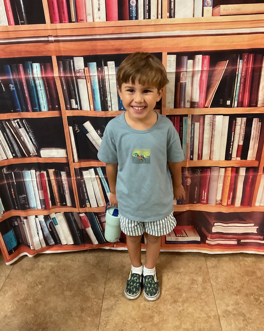 A smiling boy in a grey-blue t-shirt and striped shorts standing in front of a bookshelf backdrop on the first day of school.