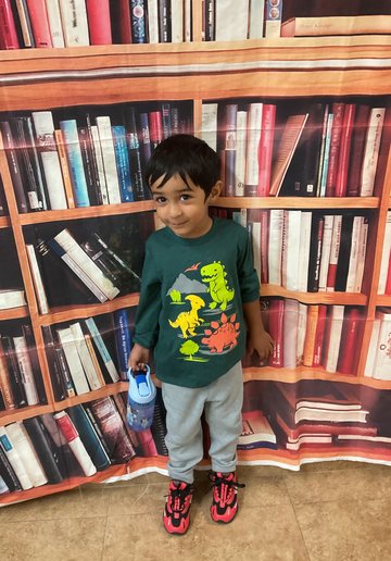A boy in a green dinosaur-print shirt holding a water bottle, in front of a bookshelf backdrop.