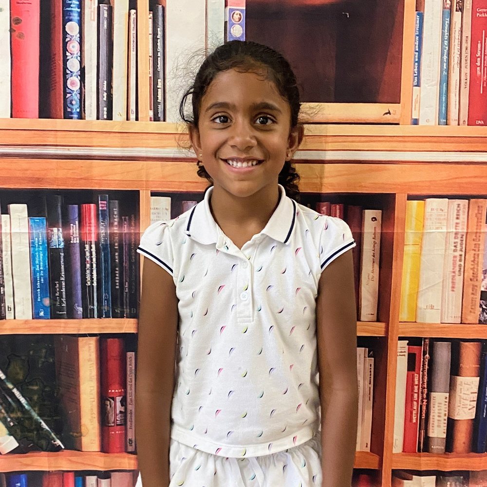 A smiling girl with dark braided hair in a white collared shirt, standing in front of a bookshelf backdrop on the first day of school.