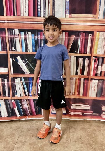 A boy in a blue t-shirt and black shorts smiling, standing in front of a bookshelf backdrop.