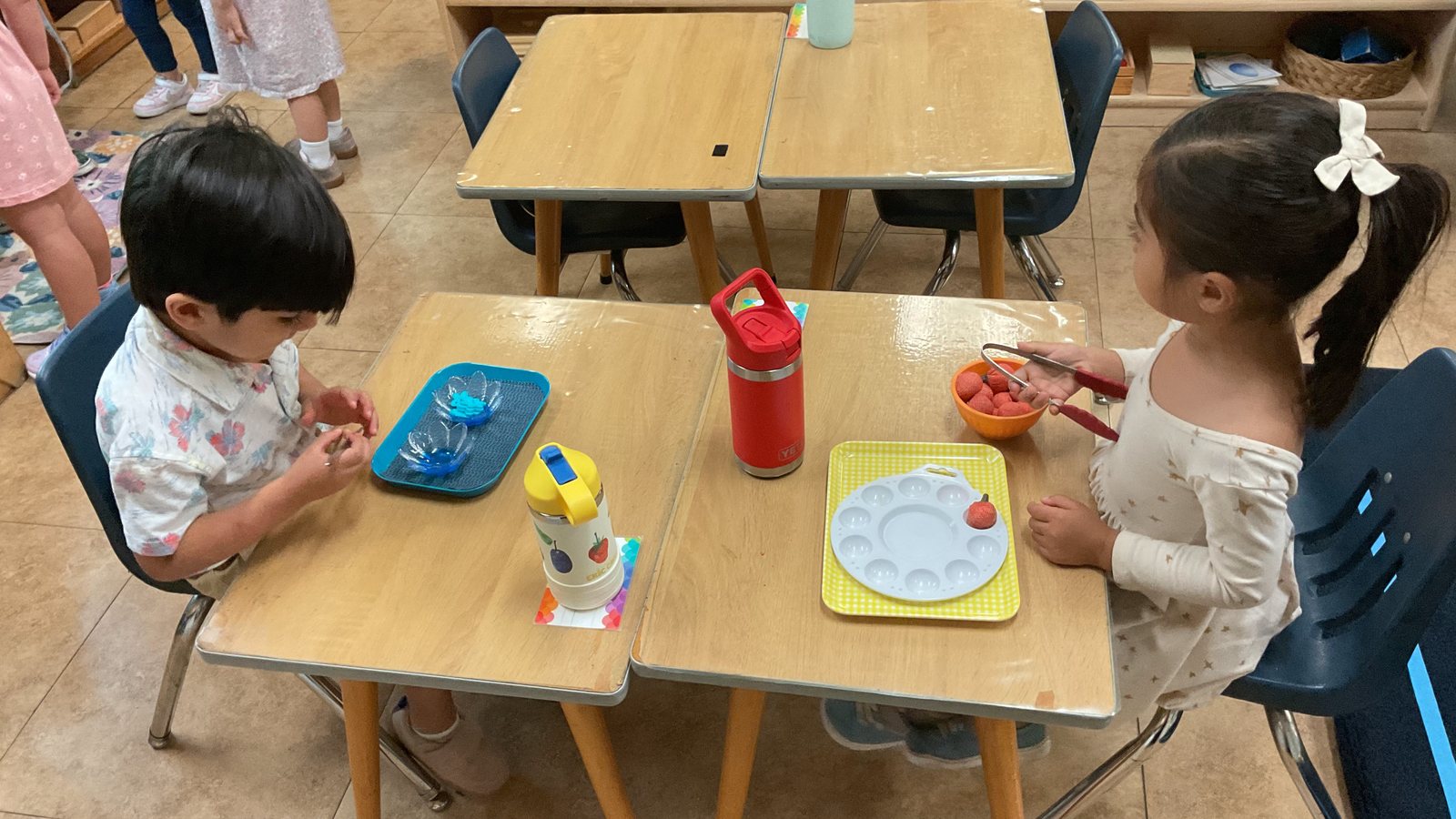 Two children at light-wood tables in a Montessori classroom, a boy in a floral shirt and a girl with a white hair bow, each working with a tray of materials.