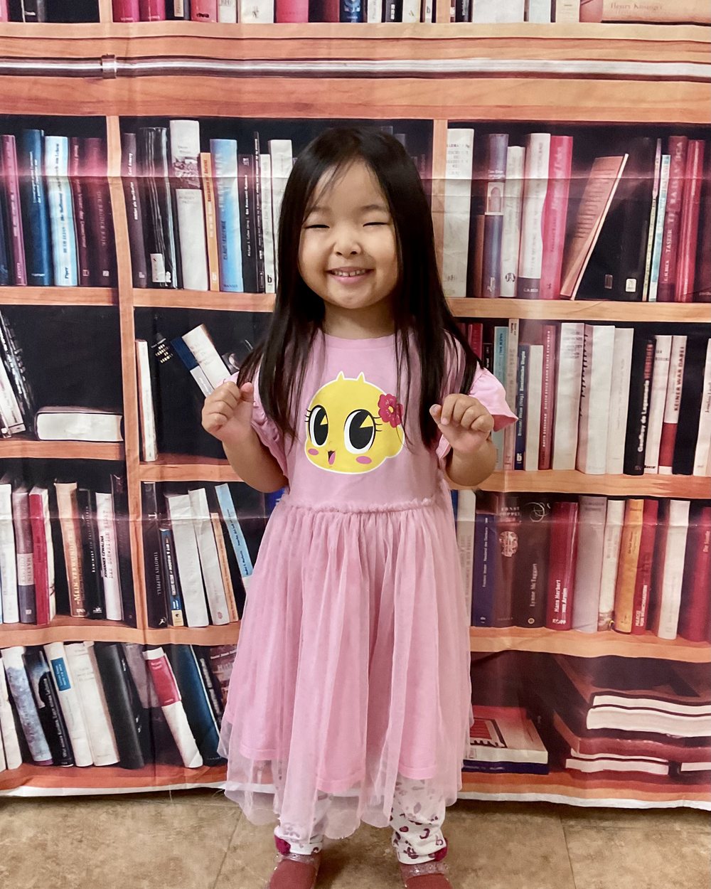 A joyful girl with long dark hair in a pink tulle dress with a yellow chick design, hands raised, on her first day of school.