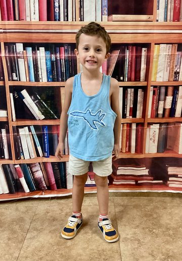 A boy in a light-blue tank top with an aeroplane graphic, standing in front of a bookshelf backdrop.