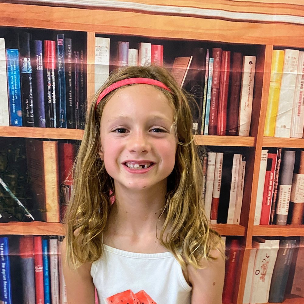 A grinning girl with long wavy hair and a red headband, missing a front tooth, on her first day of school.