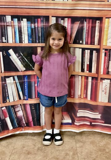 A girl in a purple ruffled top and denim shorts smiling, with a bookshelf backdrop.