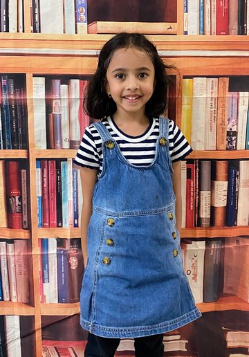 A smiling girl in a denim pinafore over a striped top, in front of a bookshelf backdrop.