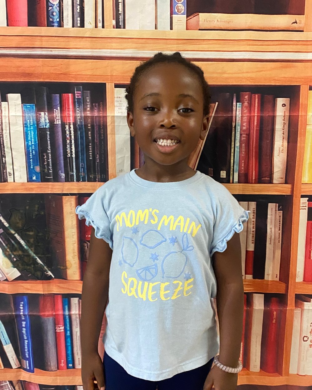 A girl with a wide smile in a light-blue ruffled shirt, standing in front of a bookshelf backdrop on the first day of school.