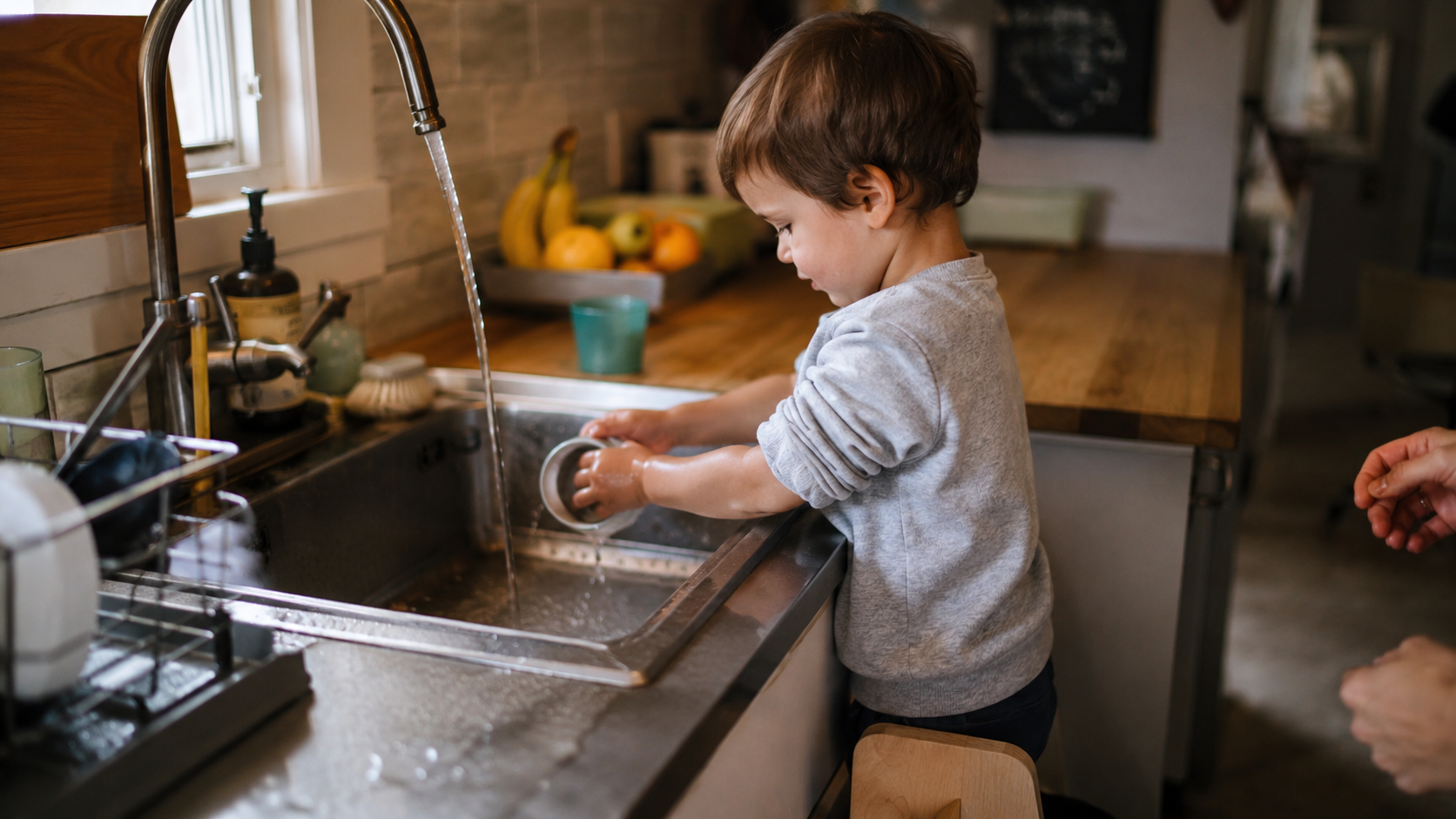 A small child standing on a low step stool washing a dish at the kitchen sink