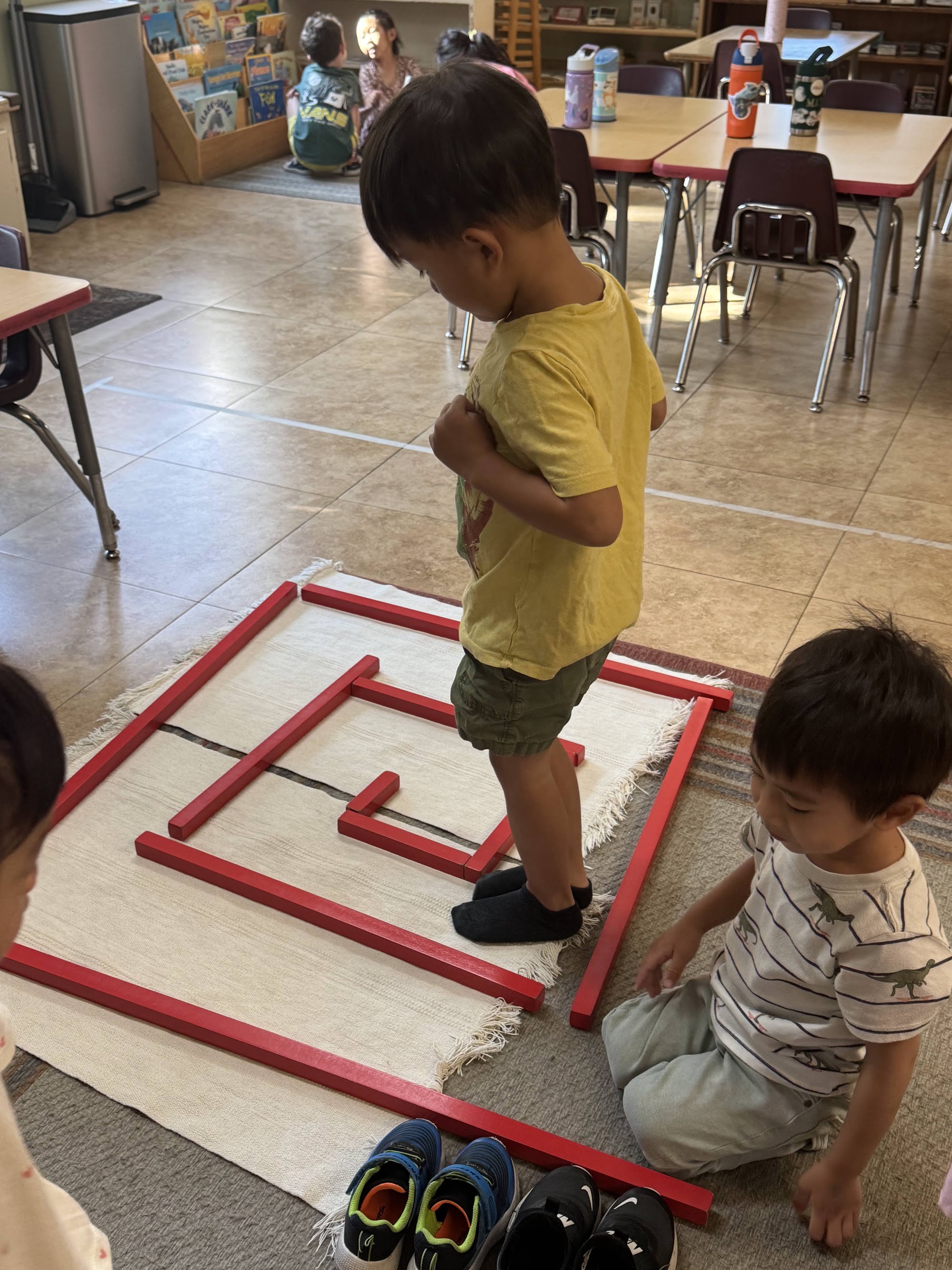 Two boys at a red-rod walking-the-line square on a work rug, one in a yellow shirt stepping along the line while the other, in a striped dinosaur shirt, kneels beside it, with classroom tables behind them.