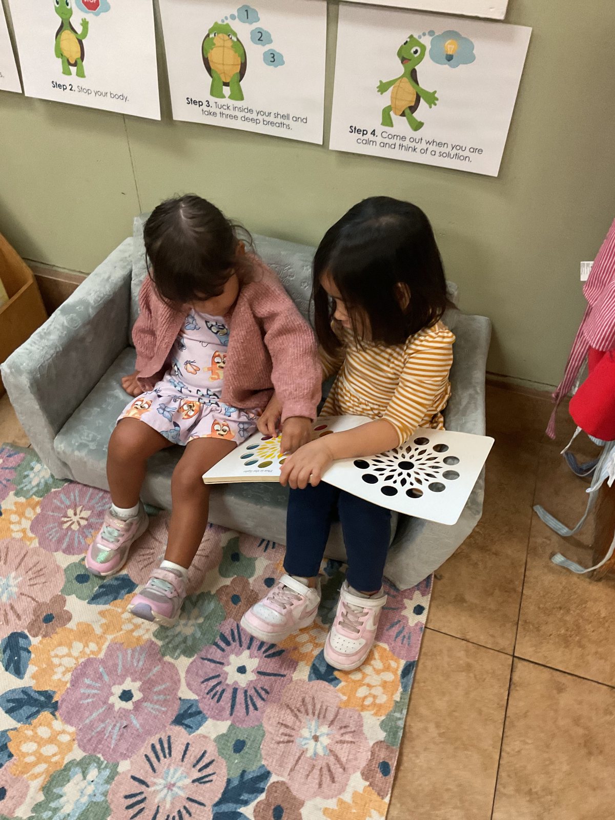 Two girls sitting close on a small grey sofa on a floral rug, sharing a book, with turtle step posters on the wall above.