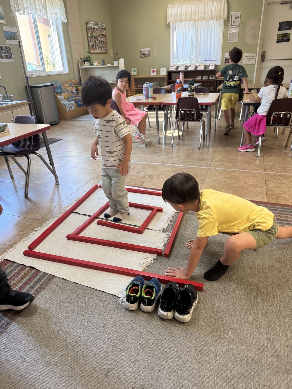 Children at work in a bright Montessori classroom, one stepping along a red line on a floor rug while another kneels beside it, other children at tables behind.