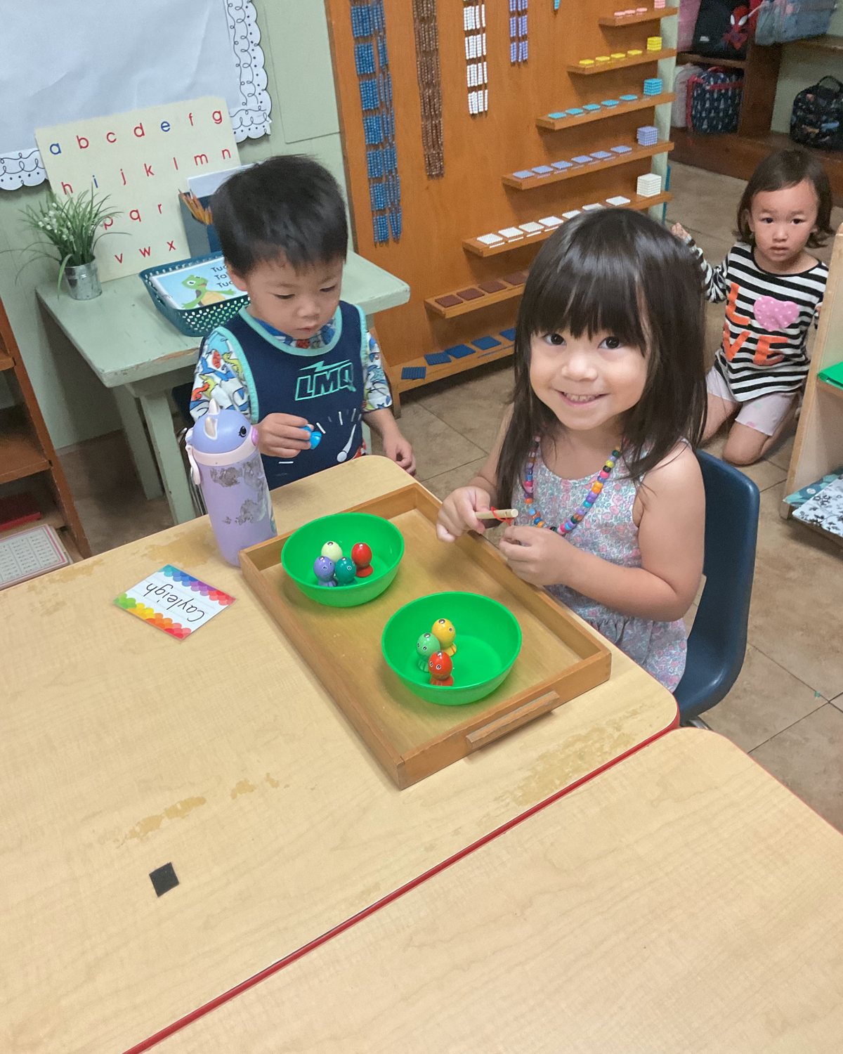 A smiling girl at a table with a wooden tray of two green bowls and small figures, a boy beside her, and Montessori geometry and alphabet materials on the wall behind.