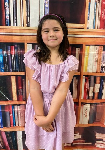 A girl with a headband in a lavender dress, arms folded, smiling slightly.