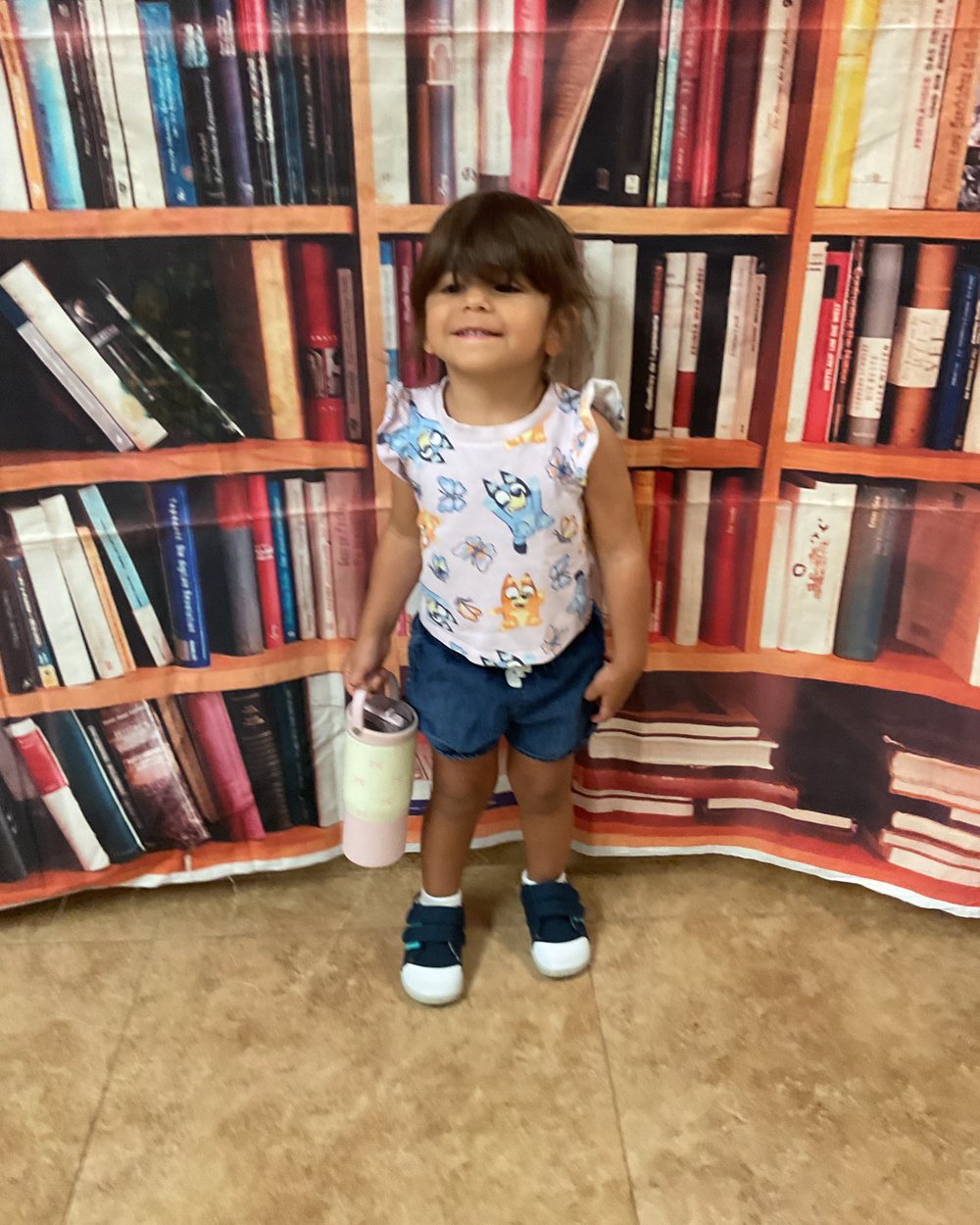 A toddler girl in a patterned top and denim shorts holding a water bottle, standing in front of a bookshelf backdrop on her first day of school.