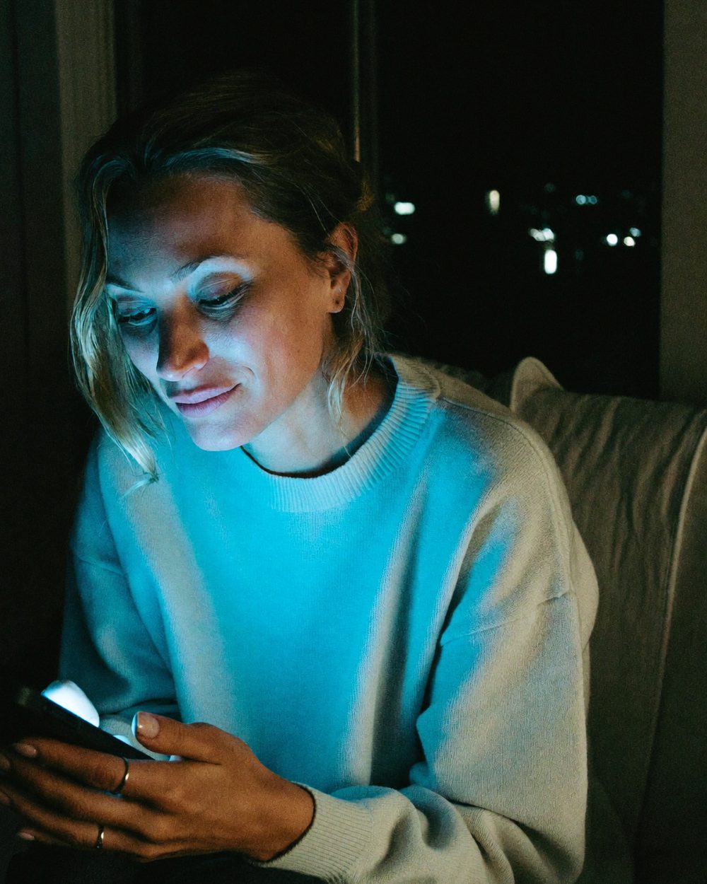 Una mujer de noche en el sofá, con la cara iluminada por el móvil, leyendo con calma.